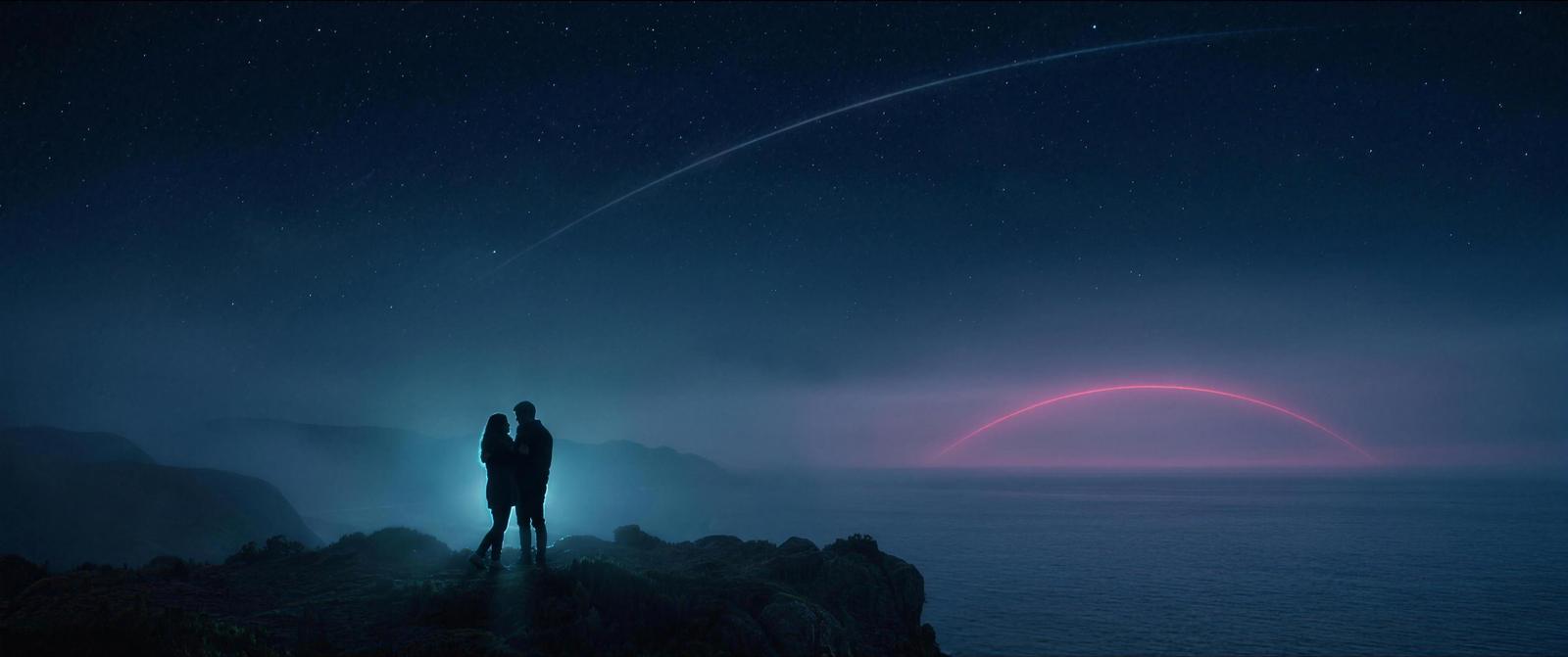 Zwei Silhouetten am Klippenrand unter dem Sternenhimmel, eine leuchtende Linie zieht über das Meer