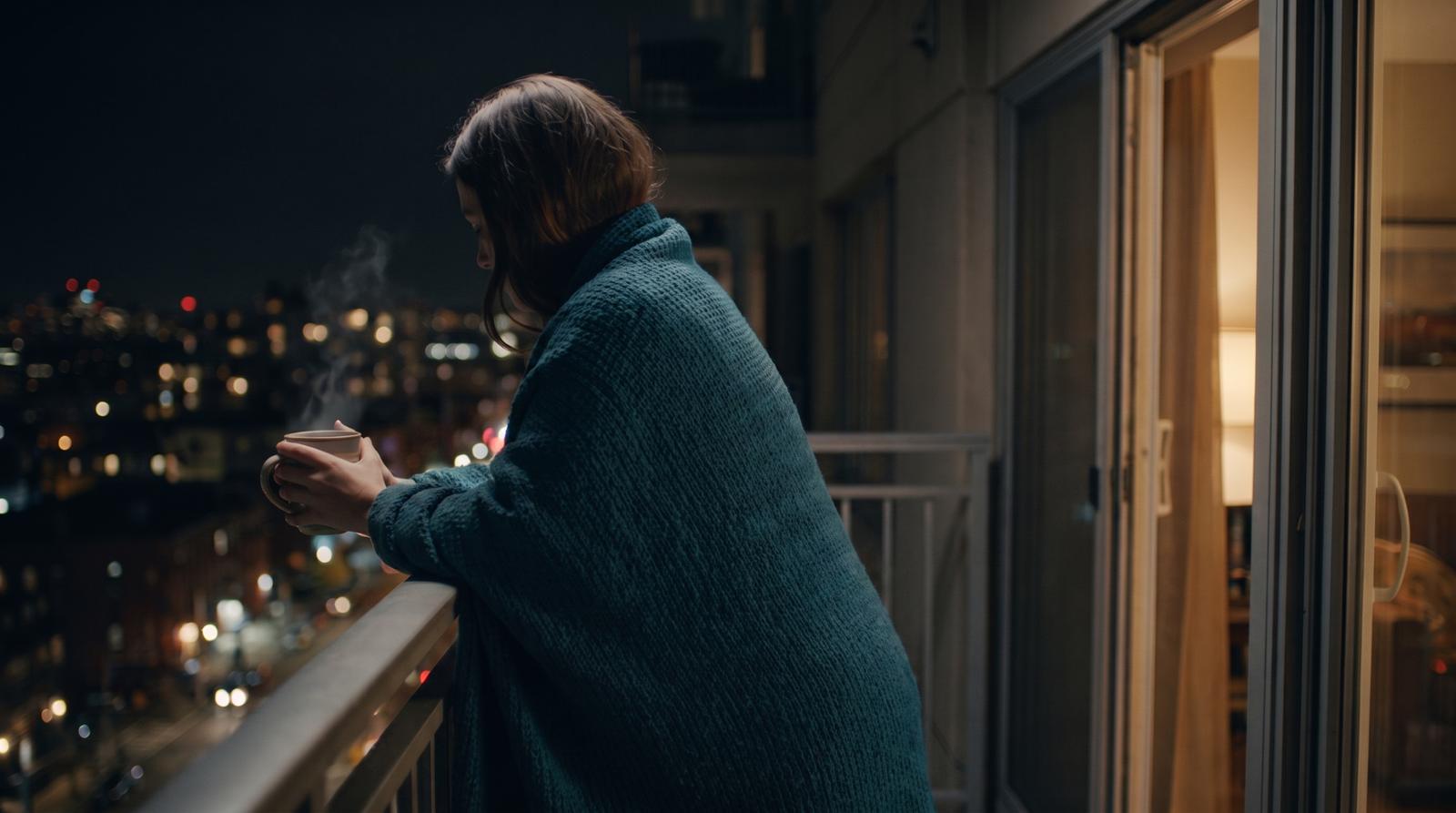 Frau in petrolblauer Strickdecke mit Tee auf einem Balkon über den Lichtern der Stadt bei Nacht – am neuen Wohnort ankommen